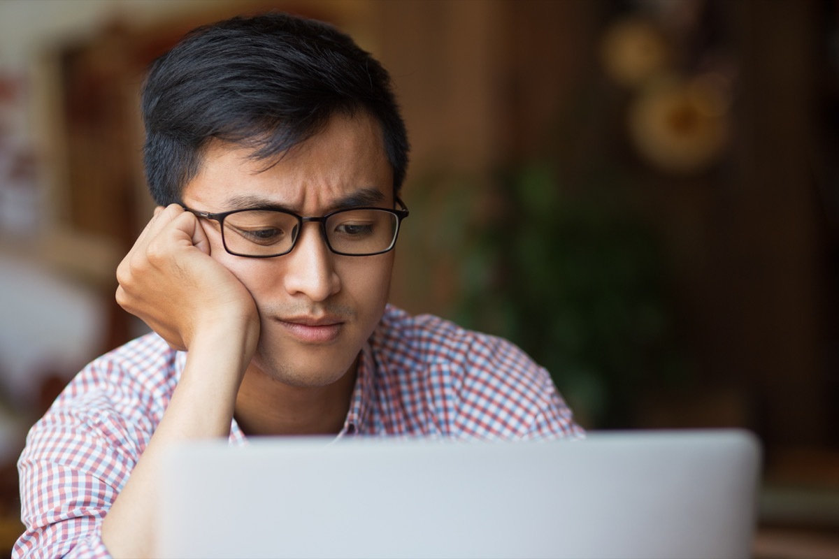 Portrait of bored young male student or businessman wearing glasses sitting at laptop leaning on arm