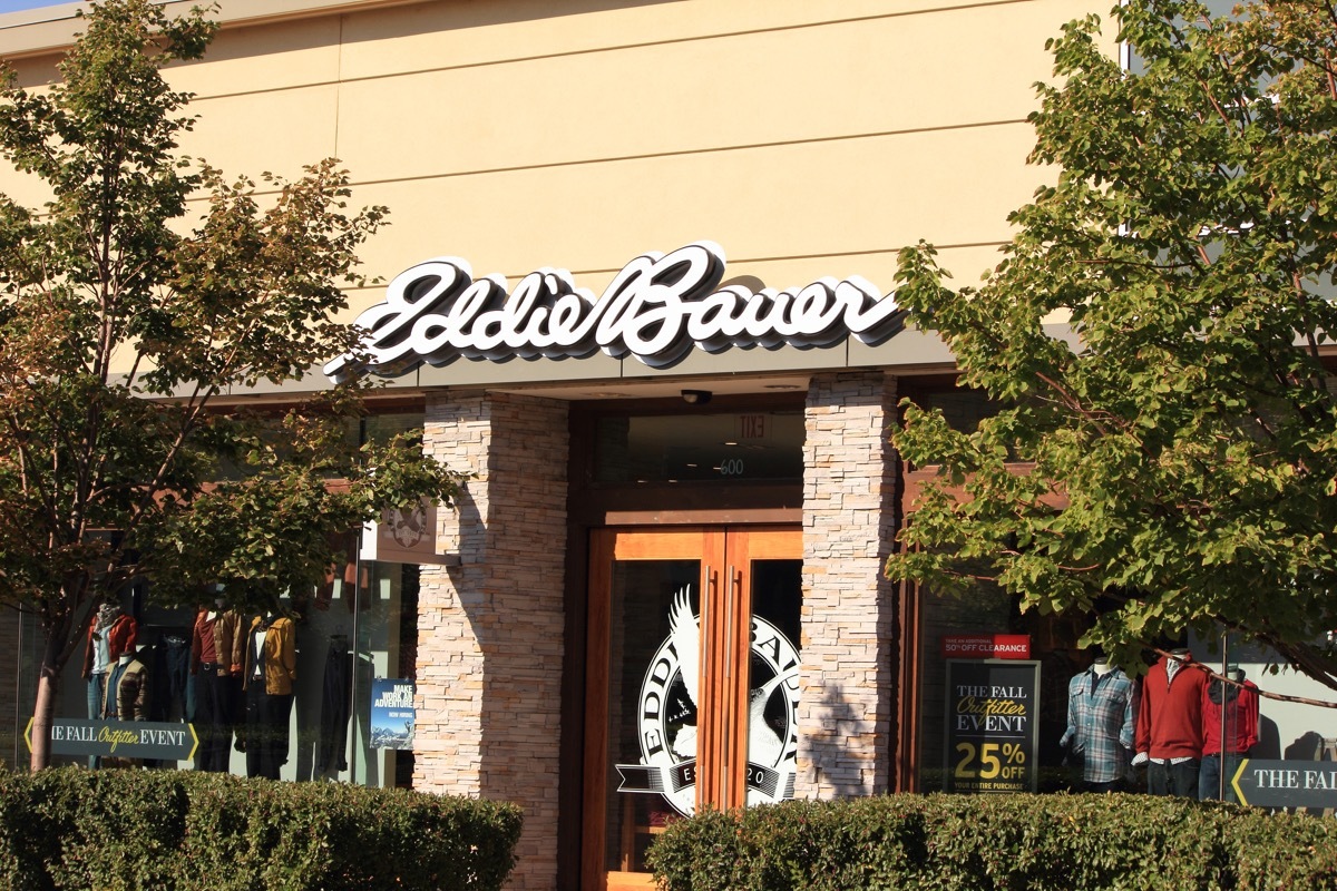 The exterior of an Eddie Bauer retail store. The large Eddie Bauer sign is in white above the entrance. The entrance door have the company logo. There are clothing displays in the store windows.