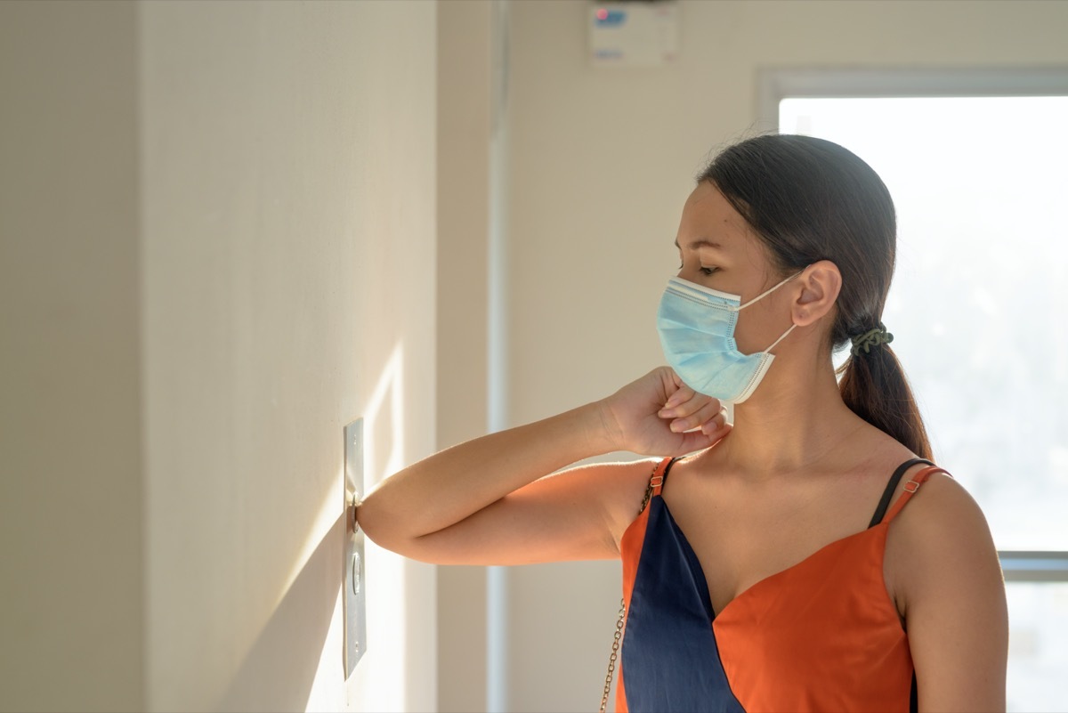 woman with mask for protection from corona virus outbreak pressing elevator door with elbow to avoid infection