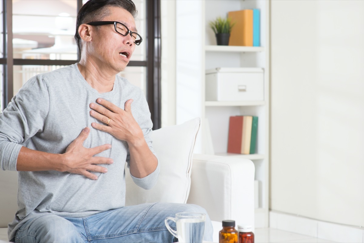 Portrait of casual 50s mature Asian man heartburn, pressing on chest with painful expression, sitting on sofa at home, medicines and water on table.