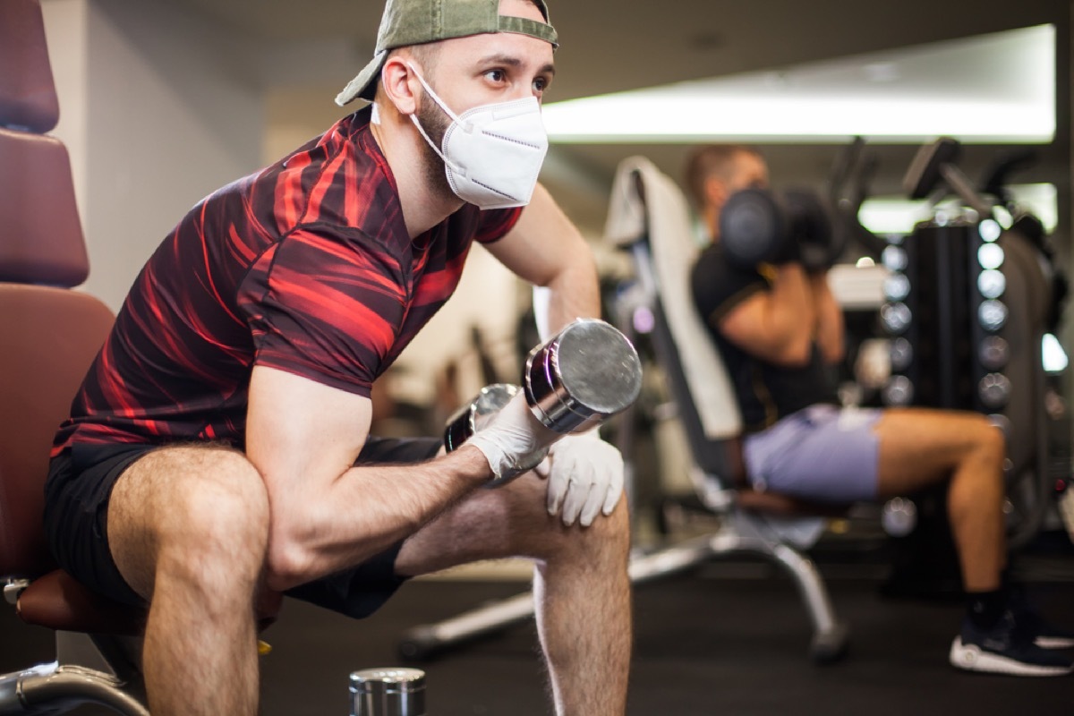 white man with face mask and gloves lifting weights at the gym